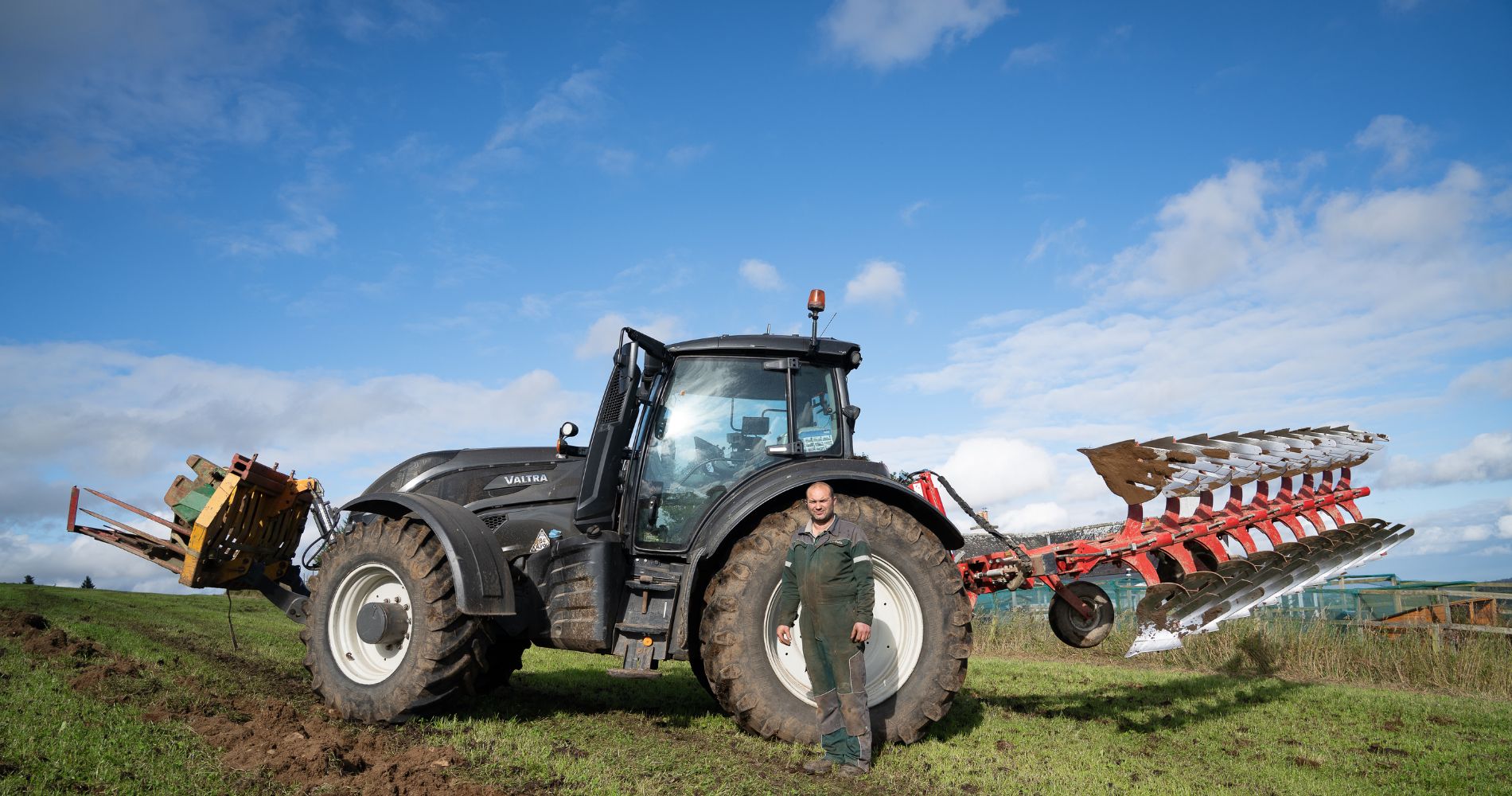 UNDO farmer partner standing beside a large black Valtra tractor with a red plow attachment on a grassy field under a blue sky with scattered clouds.