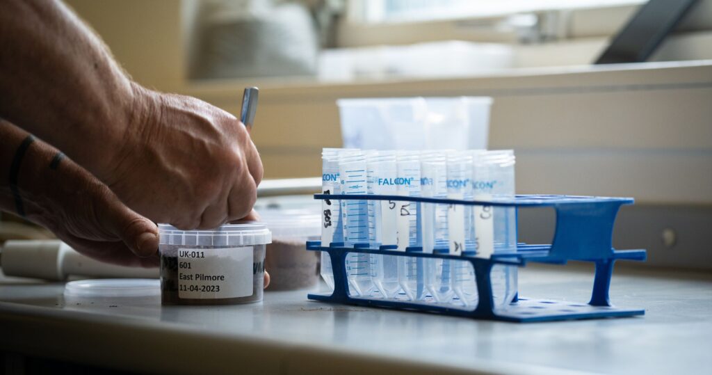 Close-up of a person's hand labeling a soil sample container alongside a rack of test tubes, representing the detailed analysis involved in carbon removal and enhanced rock weathering research.