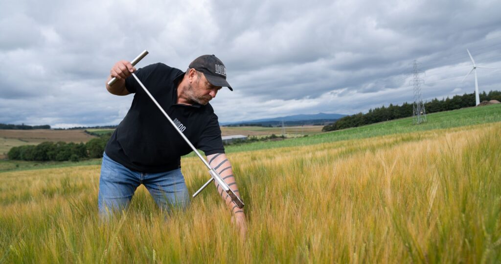 Collecting soil samples with a metal probe in a golden field, with rolling countryside and power lines visible in the background under a cloudy sky.