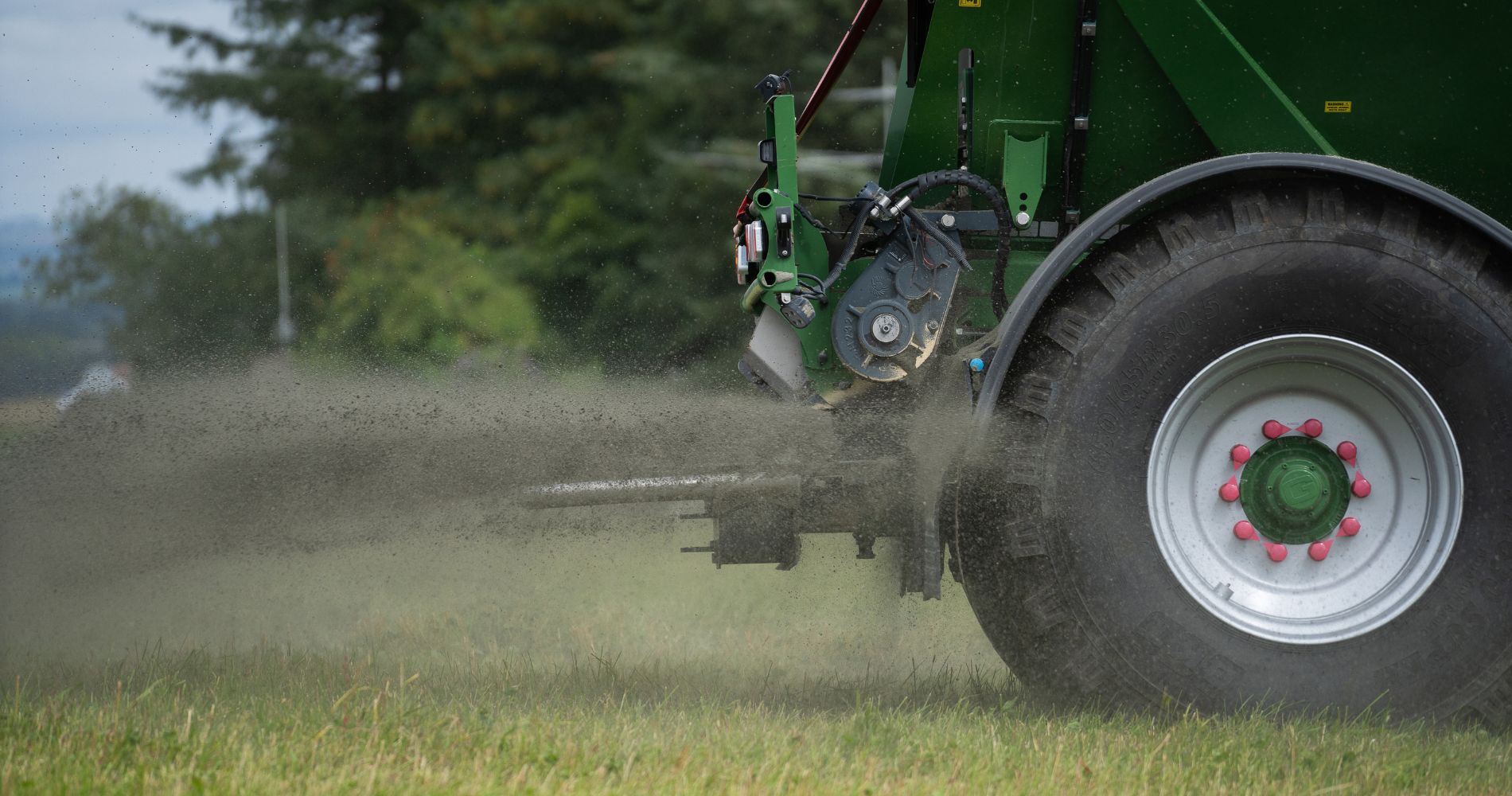 Close-up view of a green agricultural spreader attached to a tractor, dispersing fine crushed basalt rock onto a grassy field.