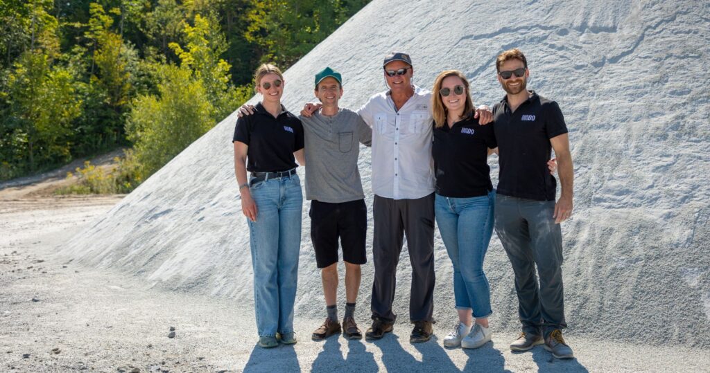 Group of five team members from UNDO and Canadian Wollastonite standing together in front of a large pile of crushed wollastonite, with trees and sunlight in the background.