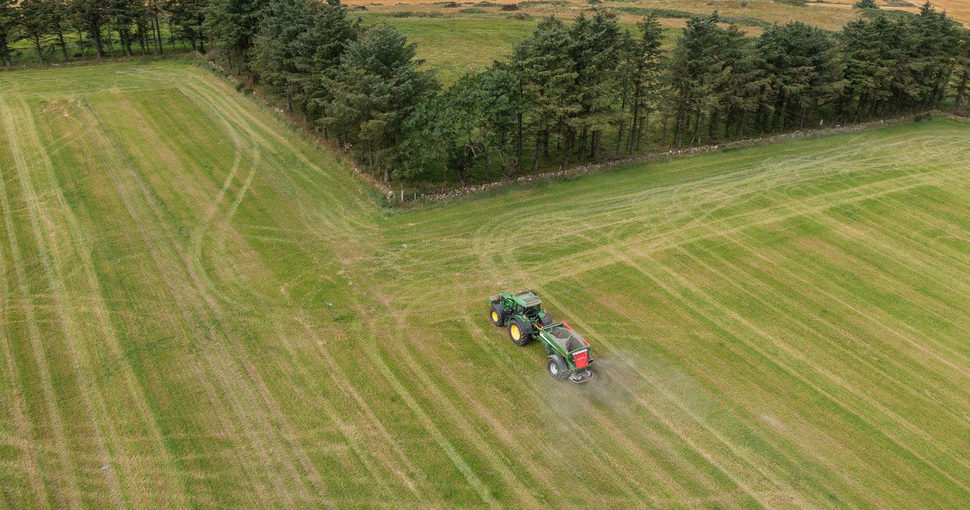 Aerial view of a green tractor spreading basalt rock over a large field, bordered by a line of tall trees.