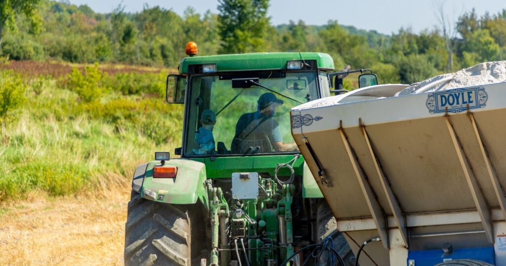 Close-up of a green tractor with a child and an adult seated inside, towing a spreader filled with crushed rock, surrounded by lush greenery and open farmland.