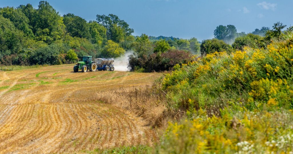 A tractor spreading crushed wollastonite across a harvested field, surrounded by green trees and wildflowers under a clear blue sky.