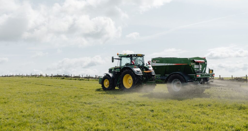 Tractor pulling a green spreader applying crushed rock across a green field under a bright, cloudy sky, showcasing a key process in enhanced rock weathering for carbon removal.